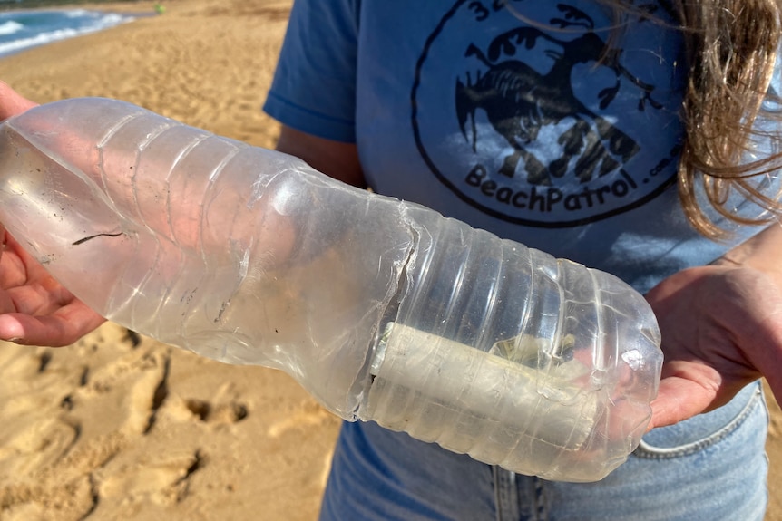 A woman's hands hold a plastic bottle, which has a note inside it. Sand is visible below the bottle and water behind it.