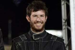 A smiling young man with dark hair, wearing a racing driver's jumpsuit.