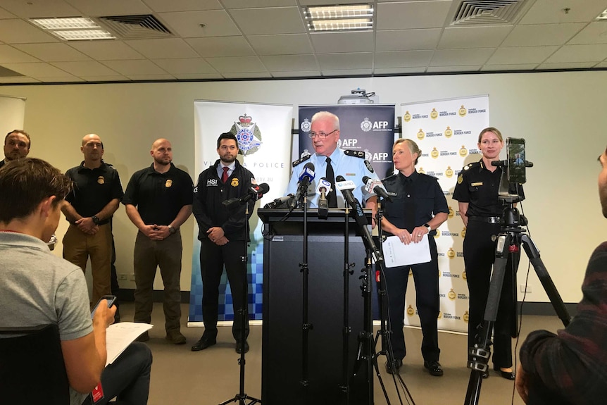 Bruce Hill wears a blue police uniform and speaking into microphones at a podium, surrounded by other officers.