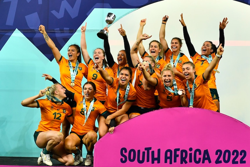 The Australian women's rugby sevens team grin and point their fingers as they wear medals and hold a cup. 
