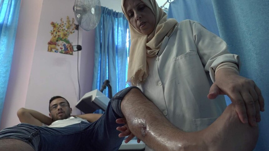 A man on a hospital bench with his leg out with a scar from a gunshot is treated by a physiotherapist.