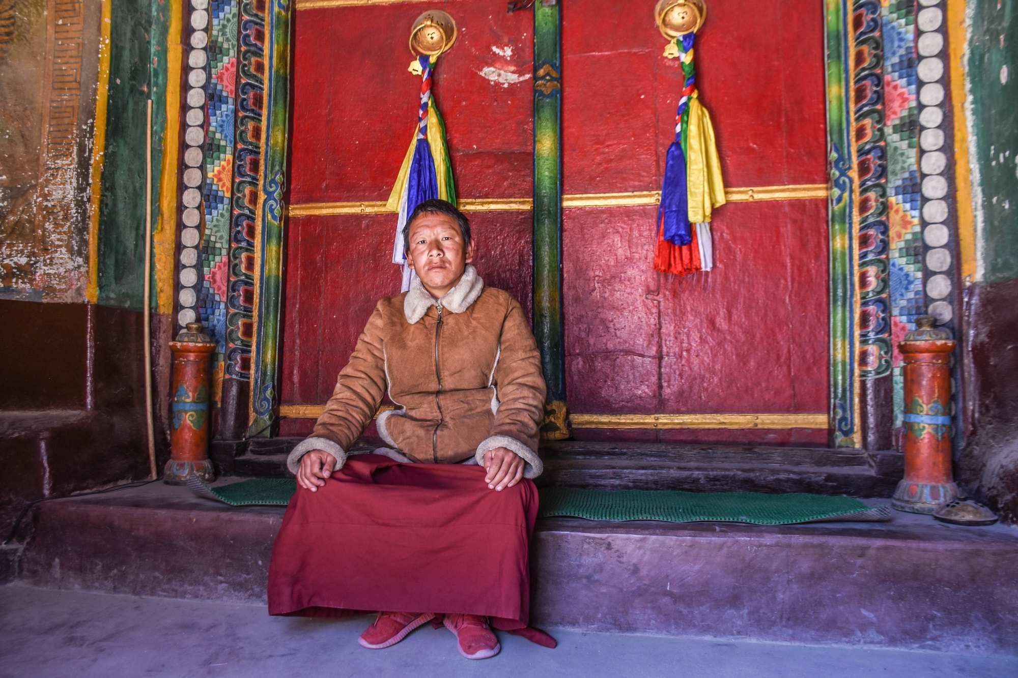 A man looks sad in a sheepskin jacket outside a colourful monastery