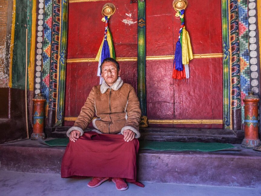 A man looks sad in a sheepskin jacket outside a colourful monastery