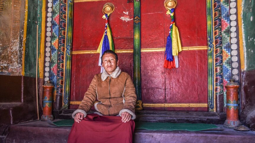 A man looks sad in a sheepskin jacket outside a colourful monastery