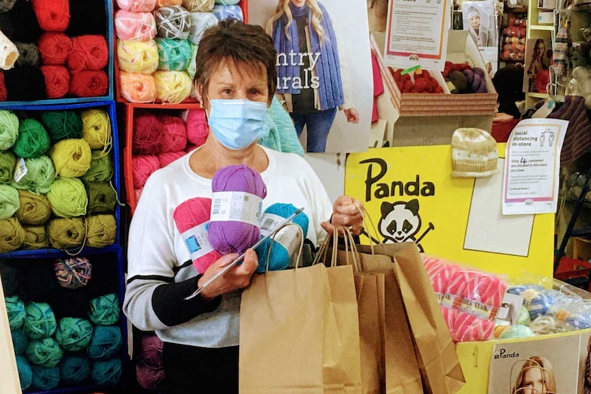 Woman with blue face mask stands in shop holding wool and bags surrounded by craft items.