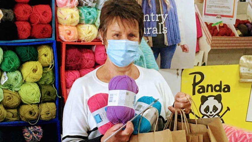 Woman with blue face mask stands in shop holding wool and bags surrounded by craft items.