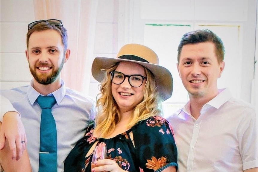 Three people, two men in collared shirts and a woman in a hat and glasses, cheerfully pose for a photo