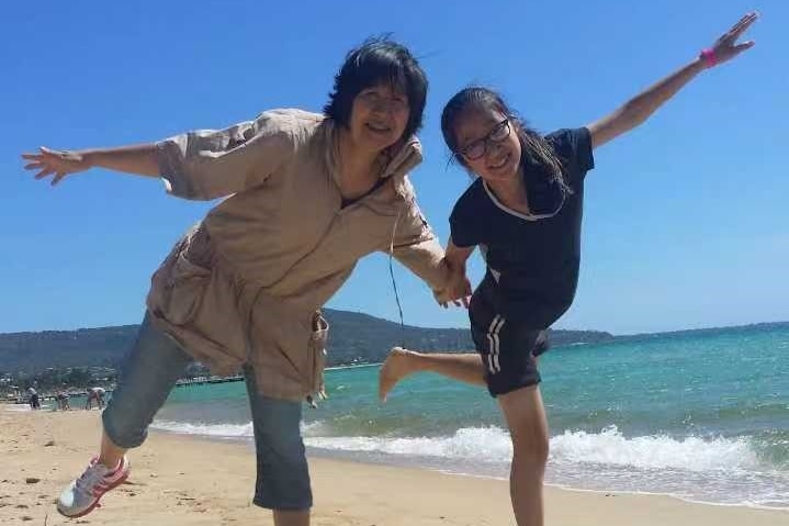 Older woman and young girl hold hands on beach and step forward into the middle of the frame.