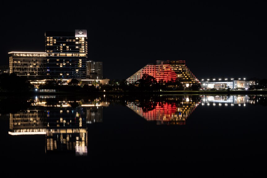 Large talk building brightly lit next to rounded lower building with both reflected in still dark water of river. 