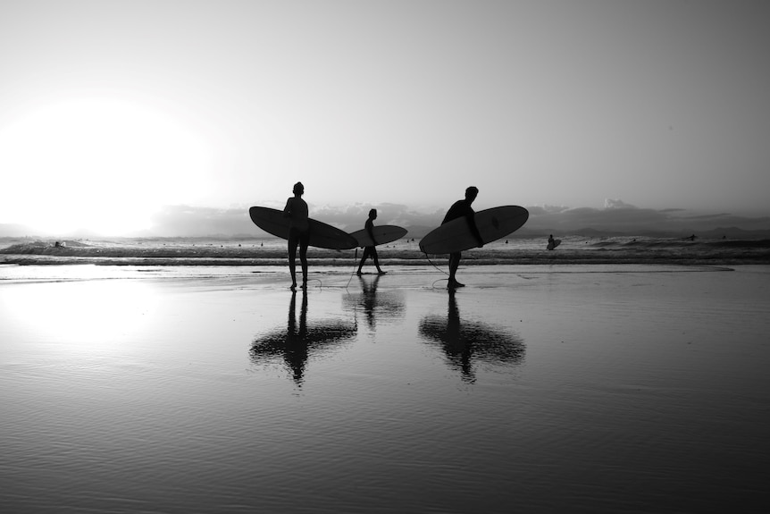 Three surfers stand on the beach in silhouette