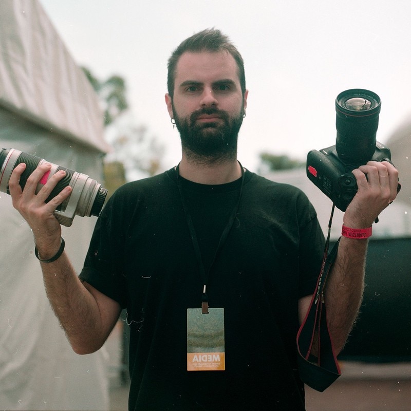 Jordan Munns stands facing the camera while holding up his camera and a long zoom lens.