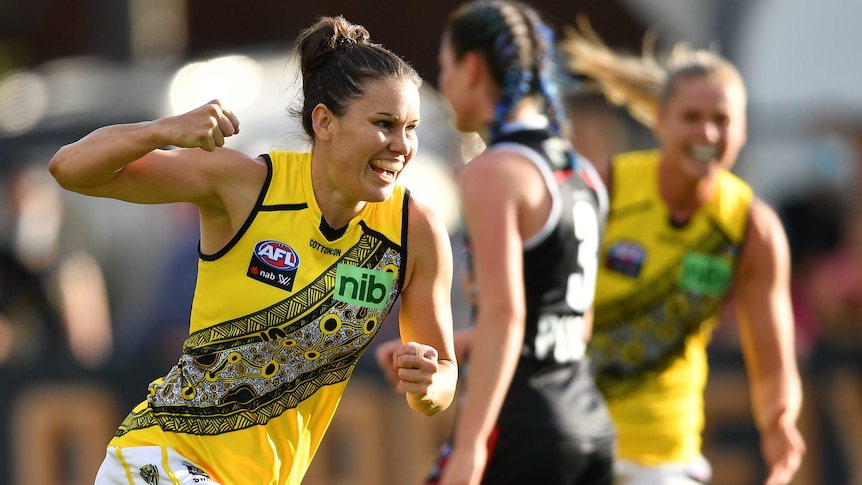 An AFLW player runs back from goal with a big smile on her face and a fist pumped in celebration.