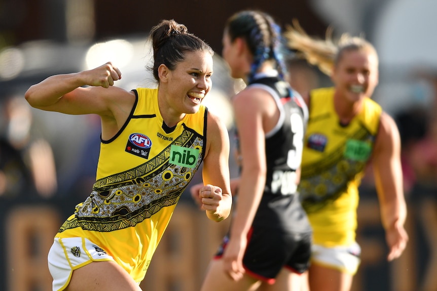 An AFLW player runs back from goal with a big smile on her face and a fist pumped in celebration.
