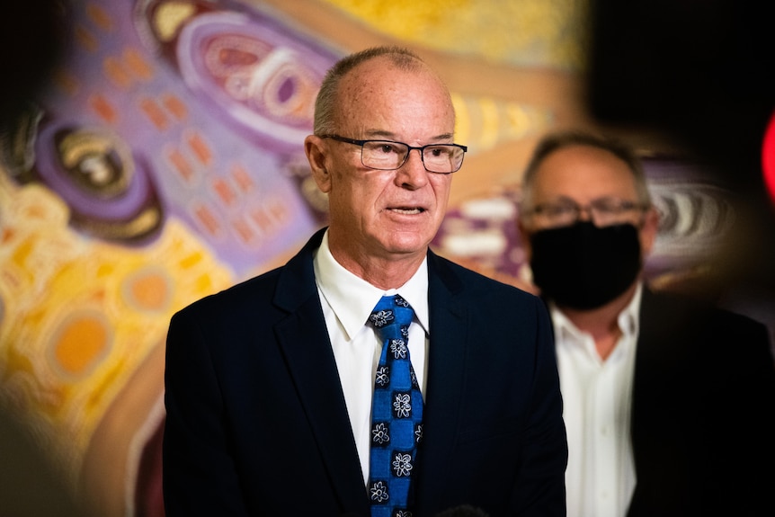 A man with glasses in a suit and dark blue tie speaks in front of a large painting.