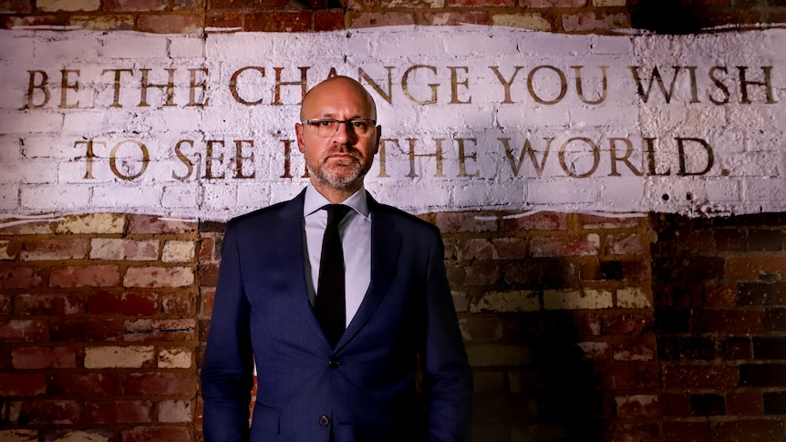 Tim Vasudeva, wearing glasses, suit and tie, stands in front of wall with the words 'Be the change you wish to see in the world'