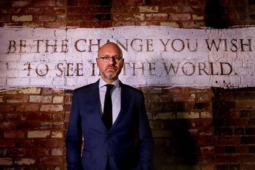 Tim Vasudeva, wearing glasses, suit and tie, stands in front of wall with the words 'Be the change you wish to see in the world'