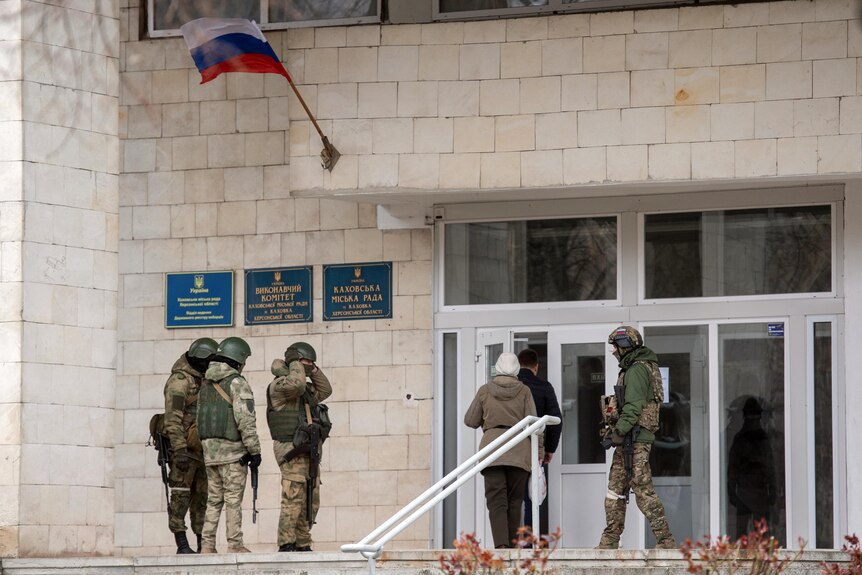 Four uniformed soldiers stand outside a building, watching civilians file through the door