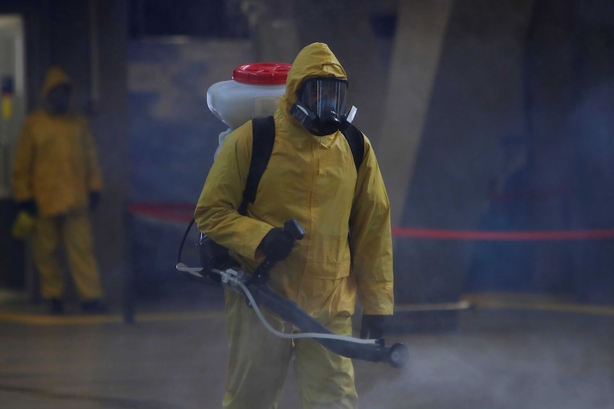 A figure in a yellow hazmat suit sprays disinfectant into the air.