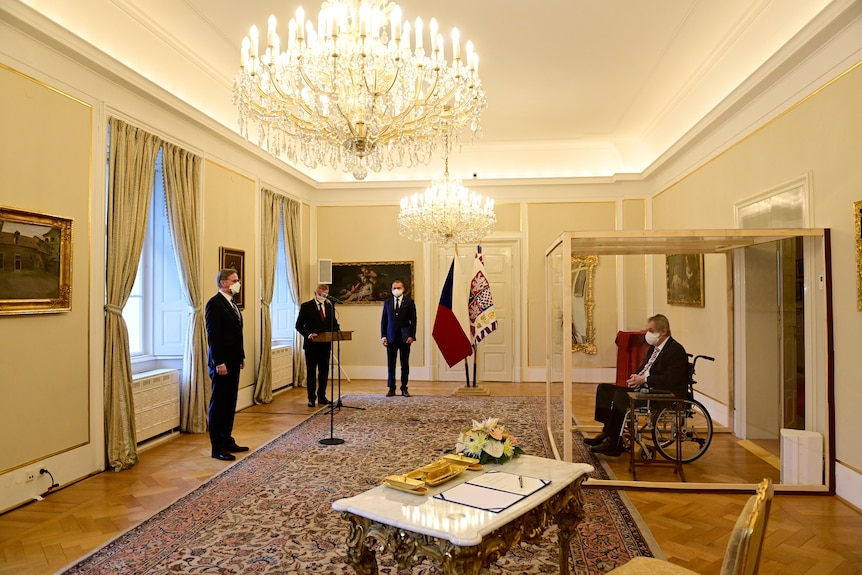 Man in a wheelchair sitting in a glass box facing three other man in suits