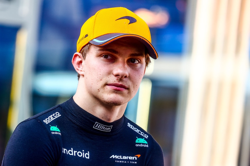 An F1 driver wearing a black shirt and orange hat looks into the distance.