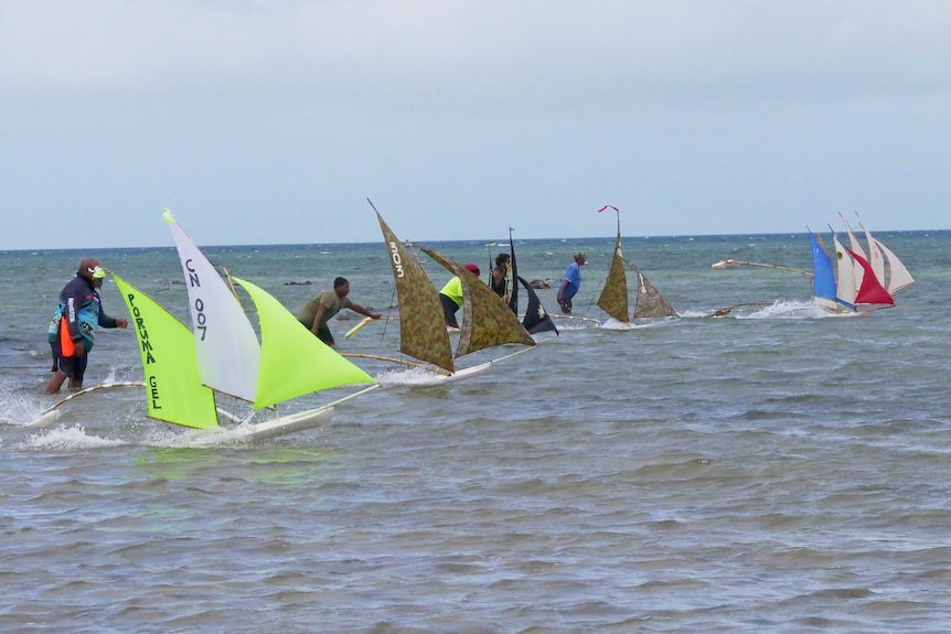 A group of men are lined up in shallow water with model boats with three sails ready to race.