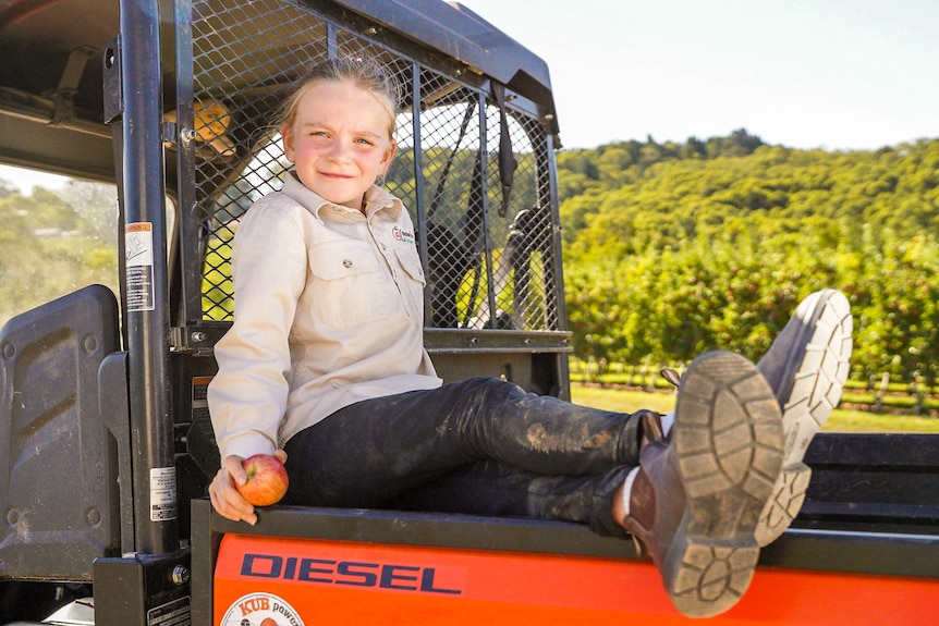 An eight-year-old girl sits comfortably on the back of a quad bike holding an apple, smiling.