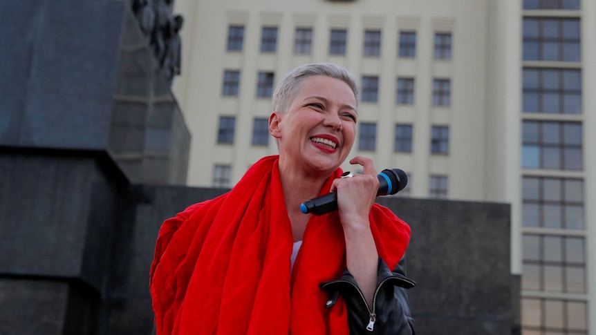 You view a woman of Eastern European descent wearing red smiling while she holds a mic in front of a Soviet-era building.