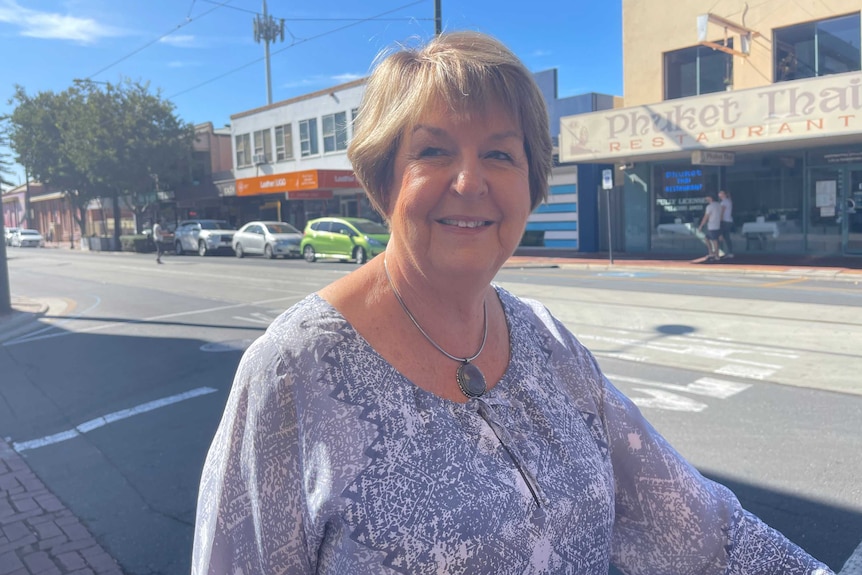 A woman with short brown hair and a grey top smiles, standing on the footpath of a main street in Adelaide.