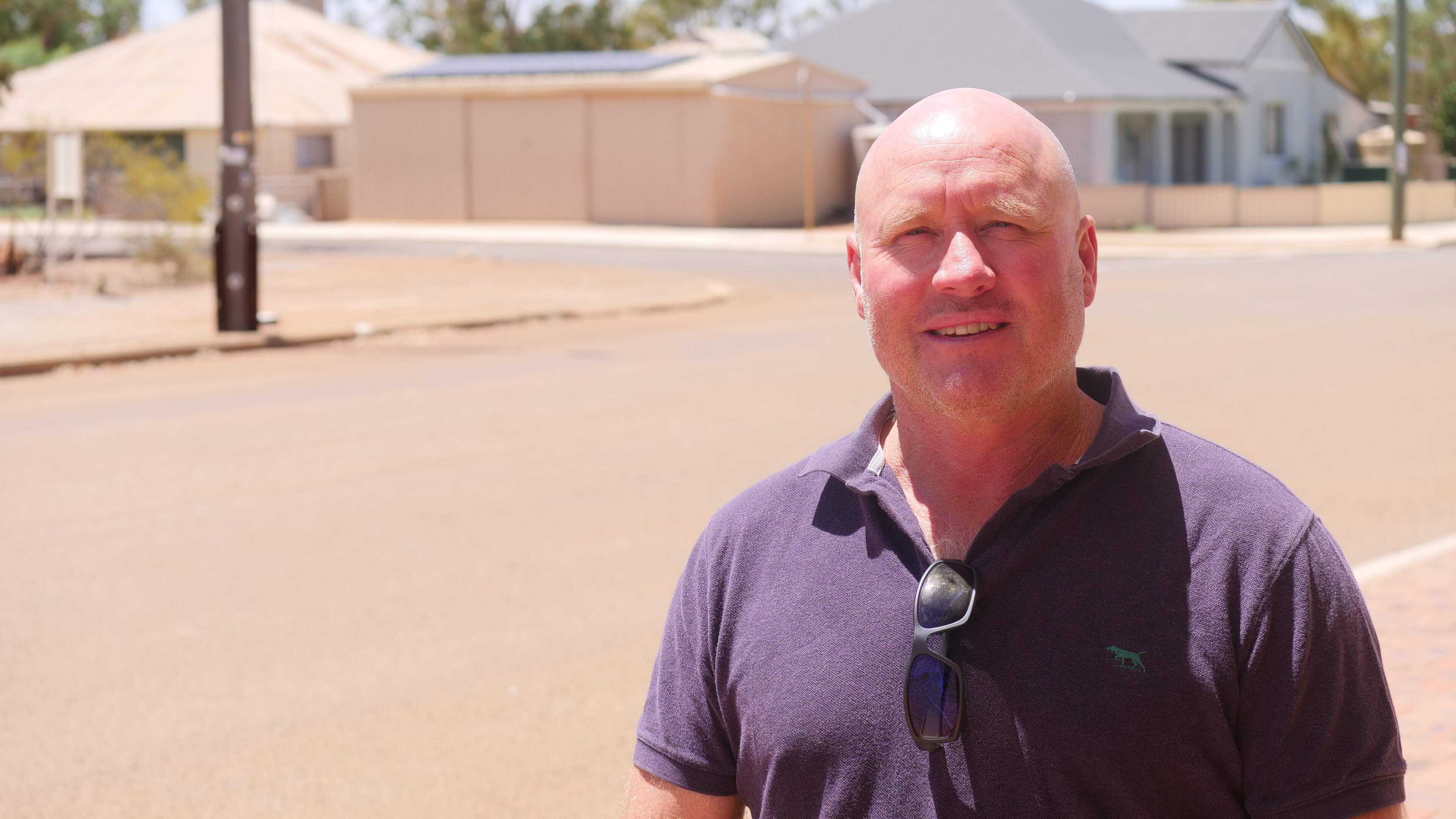 A bald man in a navy blue pollo shirt faces the camera in front of a suburban road in harsh midday sun. 