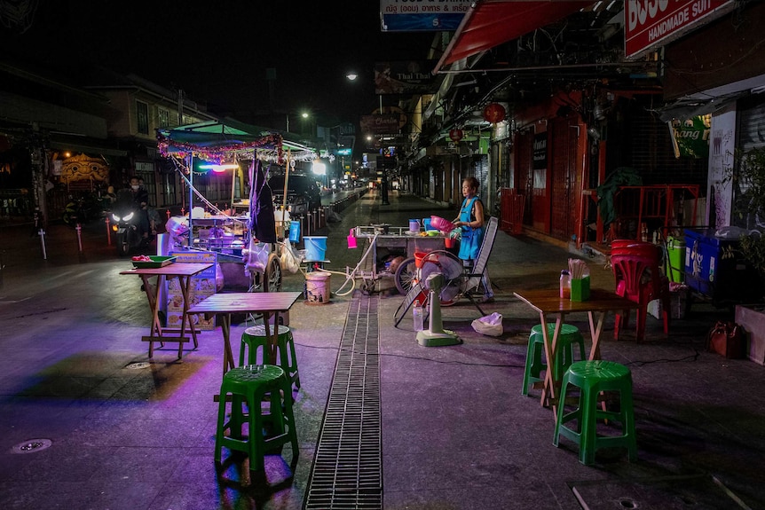 A woman in a blue apron stands alone on a dark street lit with purple light washing up at a food cart.