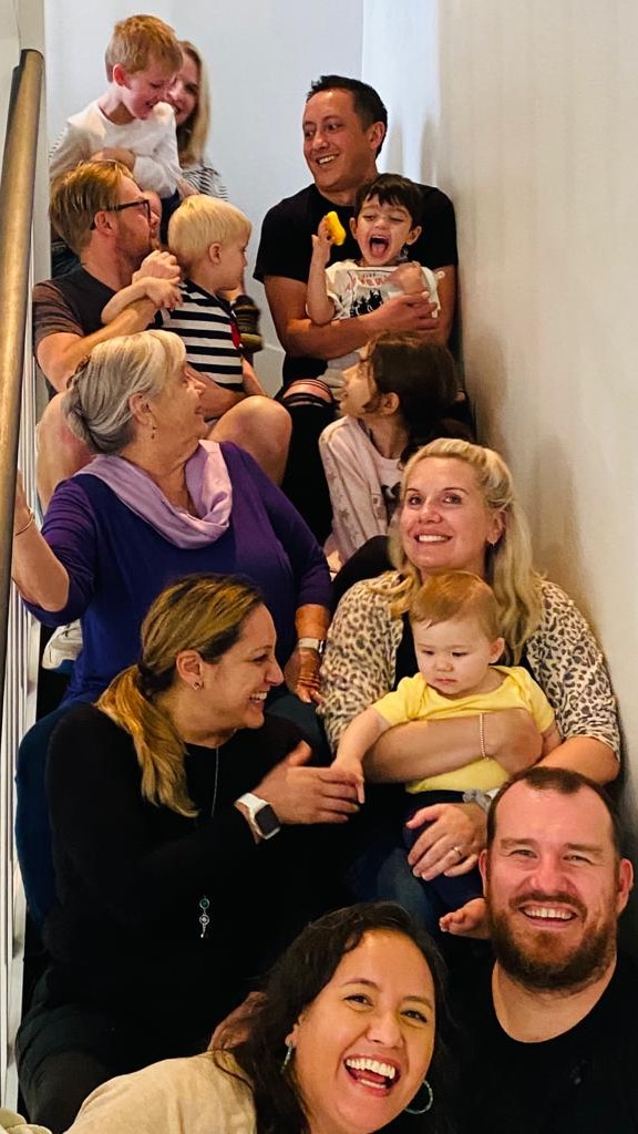 A family of 13 are sitting along a stair case, they are varying ages and are smiling and laughing.