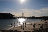 People play in the sand at Hillarys Marina, with the sun shining high overhead and boats moored in the background.