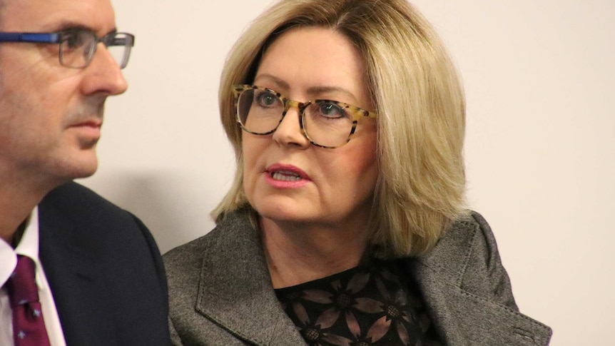 Lisa Scaffidi, in a grey jacket, black top and glasses, sits in a room next to a person in a suit.