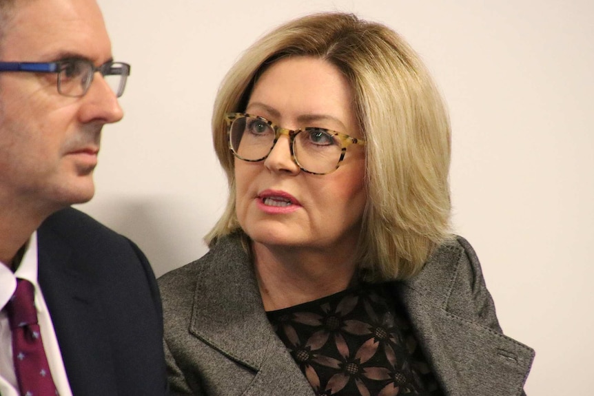 Lisa Scaffidi, in a grey jacket, black top and glasses, sits in a room next to a person in a suit.