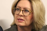 Lisa Scaffidi, in a grey jacket, black top and glasses, sits in a room next to a person in a suit.