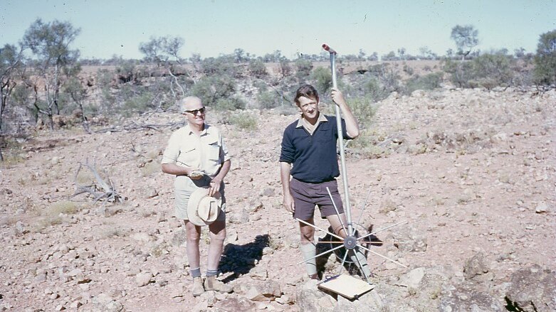 Two men stand on a rock-strewn field in 1972. One man is holding a long-handled measuring wheel.
