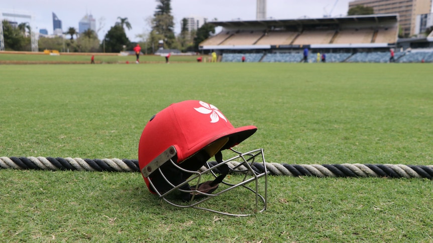Artistic shot of a red cricket helmet sitting on the turf near the oval boundary with cricketers playing in the background.