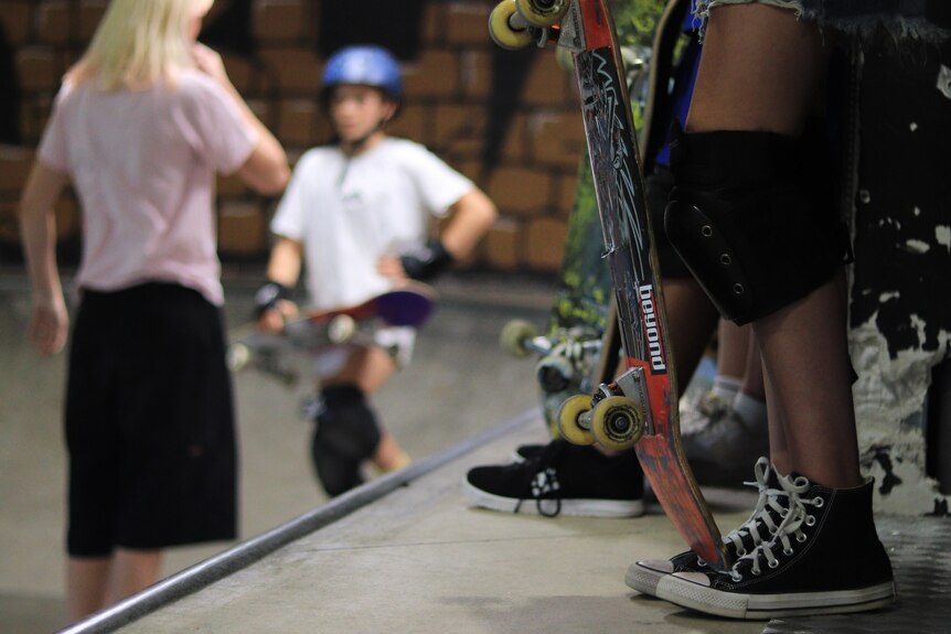 A girl's legs and a held skateboard in the foreground, the instructor talks to a student in the background. 