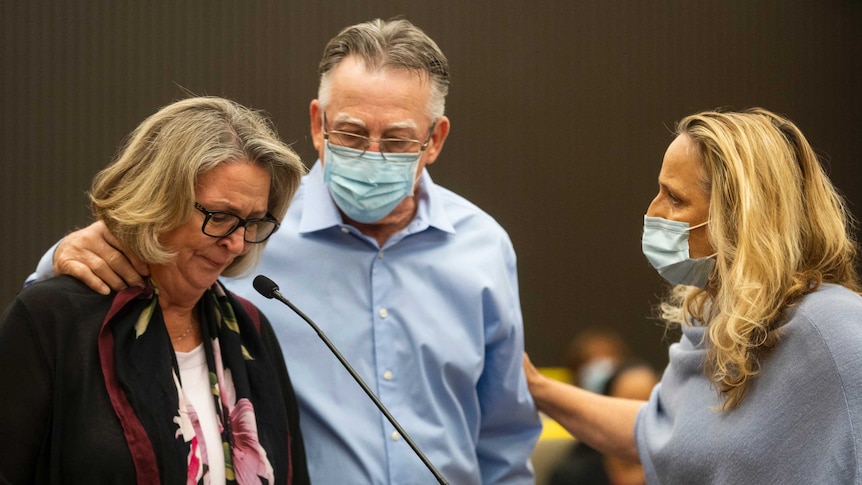 A woman cries in front of a microphone while comforted by a man and woman both in face masks
