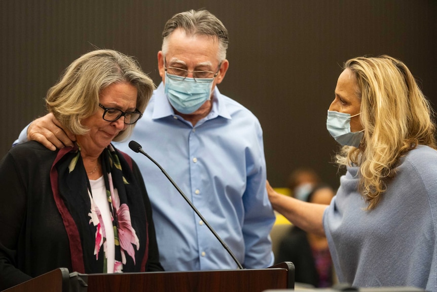 A woman cries in front of a microphone while comforted by a man and woman both in face masks
