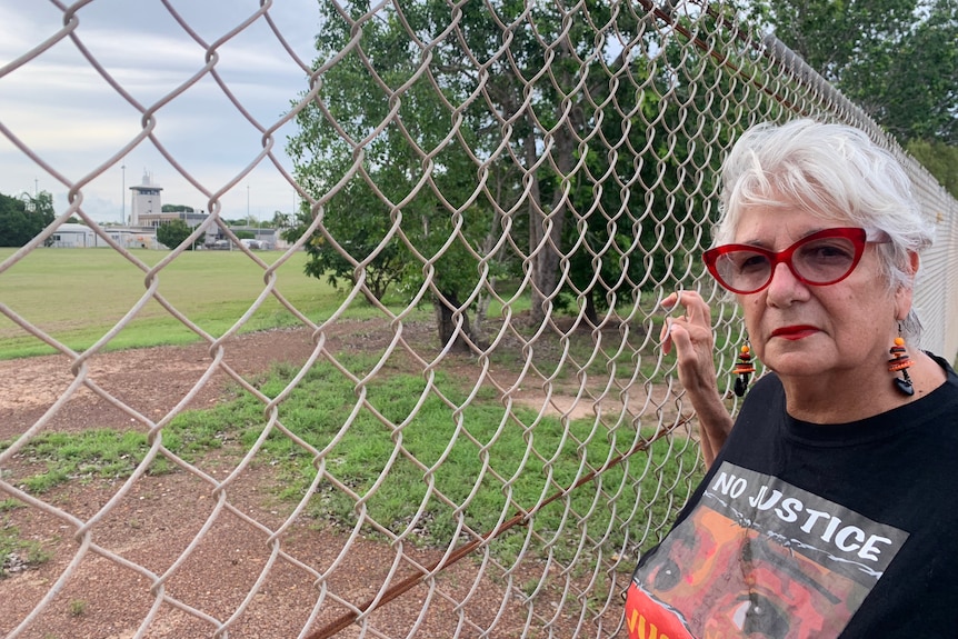 a woman wearing red glasses poses in front of mesh fence outside a detention centre