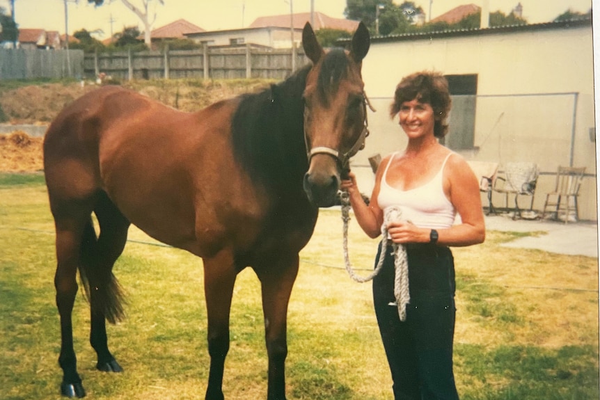 A woman with brown hair wearing a white singlet and riding pants standing alongside a brown racehorse.