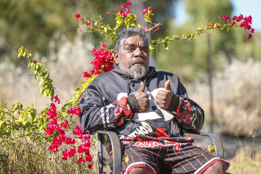 An Indigenous man poses with his thumbs up in front of a fuchsia bougainvillea plant.