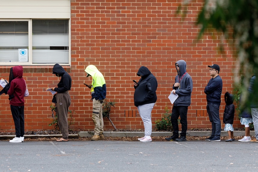 People wearing hoodies and looking at their phones as they line up alongside a brick wall.