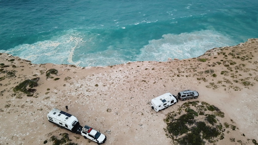 Aerial photo of two caravans attached to cars parked near the edge of a cliff, with an expanse of blue ocean below.