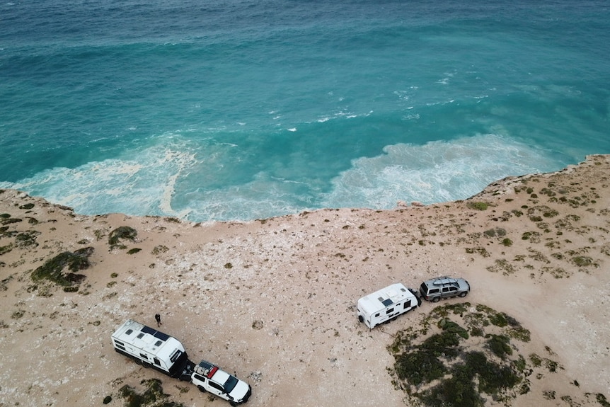 Aerial photo of two caravans attached to cars parked near the edge of a cliff, with an expanse of blue ocean below.
