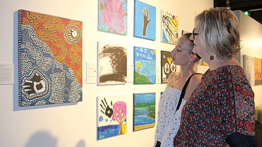 Ursula Tate and Melissa Andrew stand side-on looking at colourful dot painting inside Cairns art gallery.