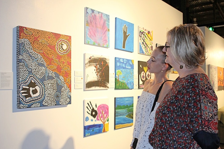 Ursula Tate and Melissa Andrew stand side-on looking at colourful dot painting inside Cairns art gallery.