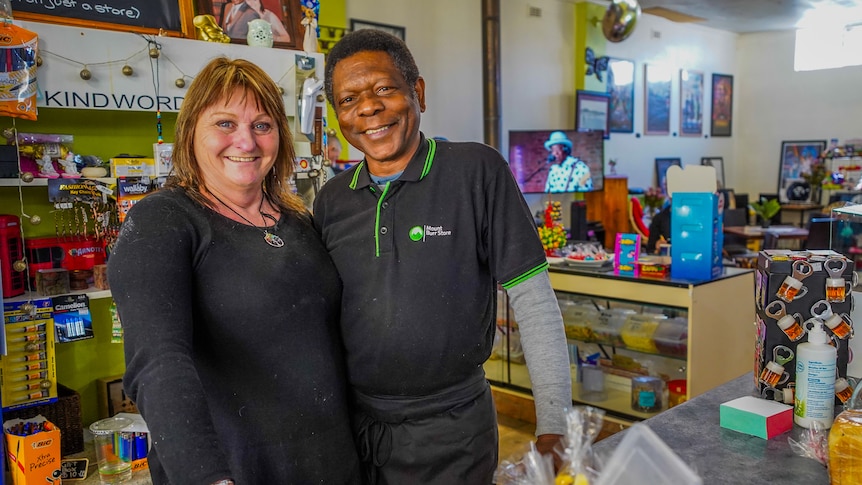A man and woman in black shirts lean against a shop counter smiling.