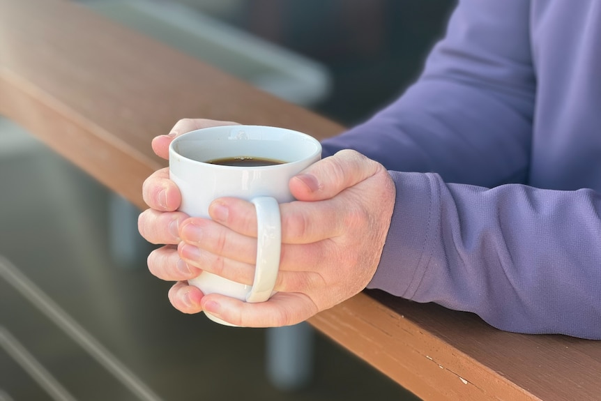 a woman wearing a purple jumper is holding a full white coffee mug, the background is blurred 
